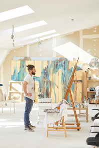 Vertical wide angle portrait of contemporary male artist looking at painting while standing by easel in sunlit art studio, copy space