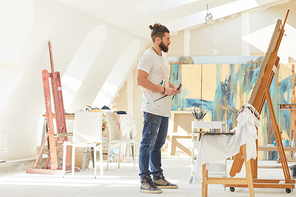 Full length portrait of contemporary male artist looking at painting while standing by easel in sunlit art studio, copy space