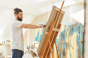 Side view portrait of contemporary male artist painting picture on easel while working in sunlit art studio, copy space