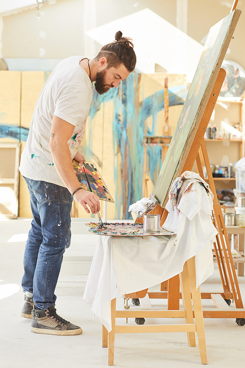 Full length portrait of contemporary bearded artist painting picture on easel while working in spacious art studio lit by sunlight