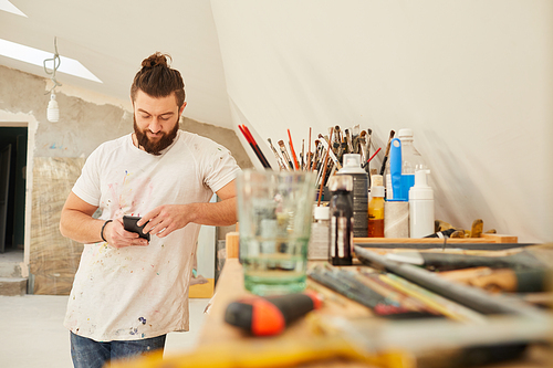 Waist up portrait of contemporary bearded artist using smartphone while standing in spacious art studio, copy space