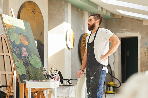 Side view portrait of adult bearded artist looking at painting on easel while standing in loft art studio, copy space