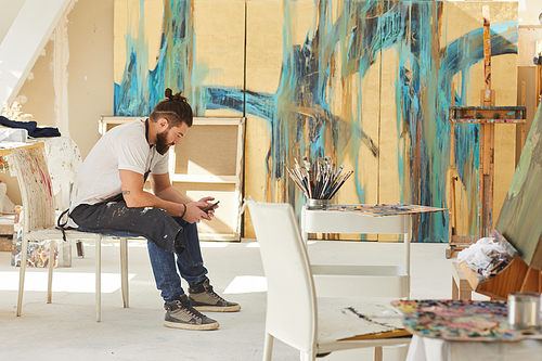 Full length side view at contemporary bearded artist using smartphone while sitting on chair in art studio with abstract paintings in background, copy space