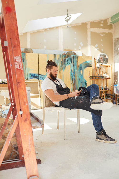 Vertical full length portrait of contemporary bearded artist using smartphone while sitting on chair in art studio with abstract paintings in background, copy space
