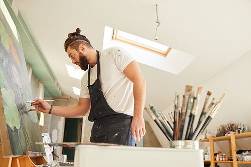 Side view portrait of contemporary bearded artist painting picture on easel with great focus and attention while standing in spacious attic art studio, copy space