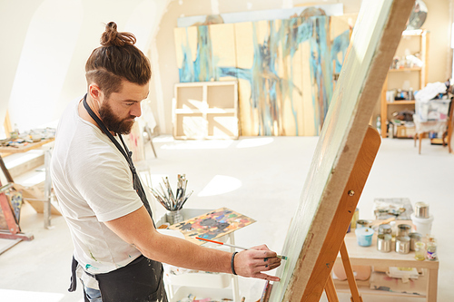 High angle portrait of contemporary bearded artist painting picture on easel with great focus and attention while standing in spacious art studio lit by sunlight, copy space