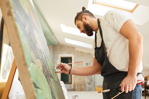 Low angle side view at adult contemporary artist painting picture on easel while standing in spacious art studio lit by sunlight