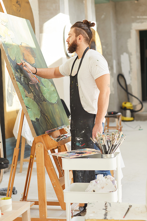 Vertical full length portrait of contemporary bearded artist painting picture on easel while working in art studio lit by sunlight