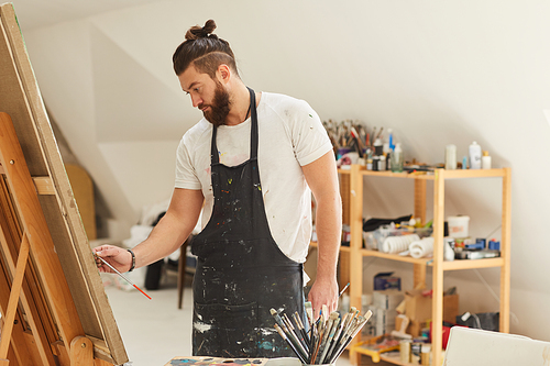 Waist up portrait of contemporary bearded artist painting picture on easel while working in spacious art studio, copy space