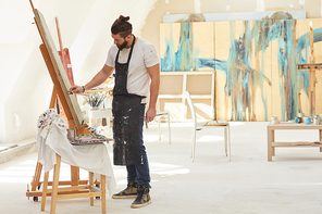 Wide angle side view at contemporary bearded artist painting picture on easel while working in spacious art studio lit by sunlight, copy space