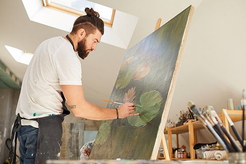 Side view portrait of talented male artist painting picture on easel while working in spacious art studio lit by sunlight, copy space