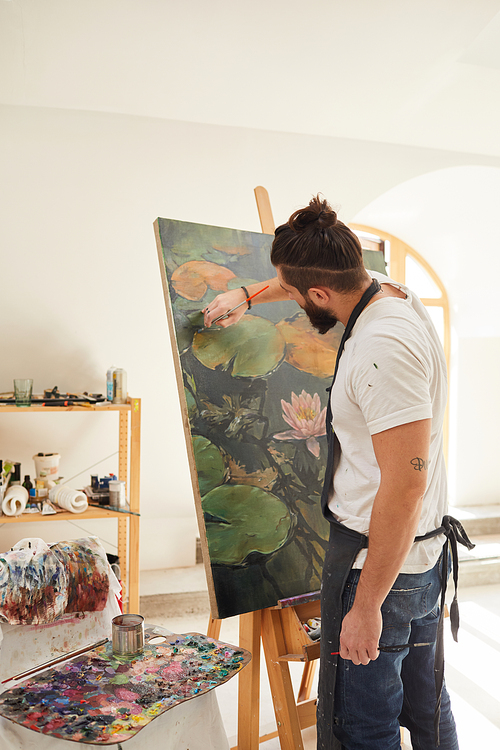 Vertical back view portrait of talented male artist painting picture on easel while working in spacious art studio lit by sunlight, copy space