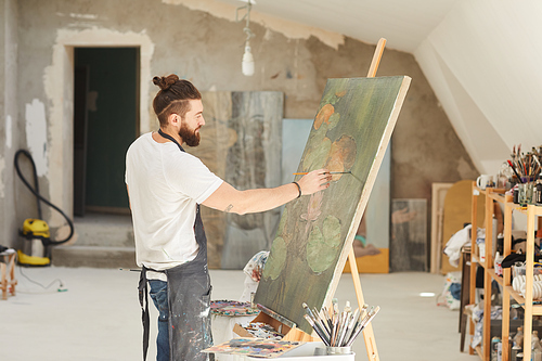 Side view portrait of talented male artist painting picture on easel while working in spacious art studio lit by sunlight, copy space