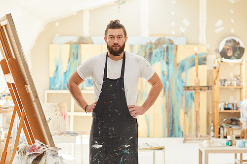 Portrait of mature bearded artist wearing apron and posing confidently  while standing in spacious art studio lit by sunlight, copy space