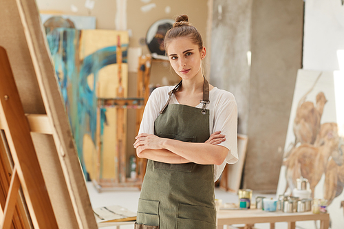 Waist up portrait of young female artist  while standing with arms crossed next to easel in art studio, copy space