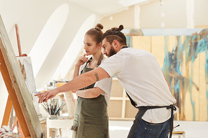 Side view portrait of contemporary young couple painting picture together while standing by easel in spacious art studio lit by sunlight, copy space