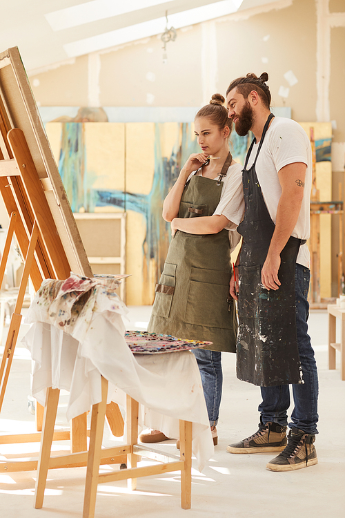 Vertical full length portrait of contemporary couple painting picture together while standing by easel in art studio, copy space