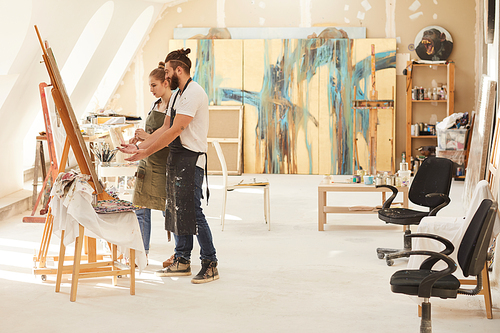 Wide angle side view at creative young couple painting picture together while standing by easel in spacious art studio lit by sunlight, copy space