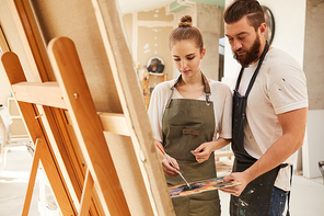 Warm toned portrait of creative couple painting picture together while standing by easel and holding palette, copy space