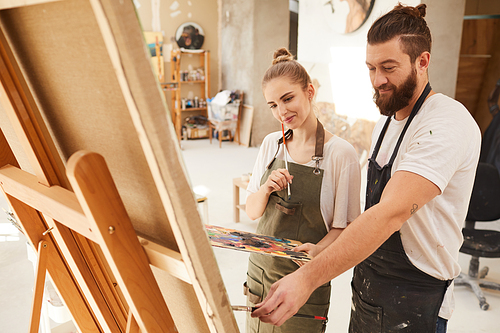 Warm toned portrait of creative couple painting picture together and smiling happily while standing by easel in sunlit art studio, copy space