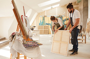 Full length portrait of two contemporary artists wrapping painting for shipping while standing in spacious art studio lit by sunlight, copy space