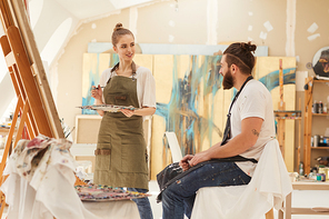 Portrait of young woman painting picture on easel while working in sunlit art studio with bearded man sitting on chair and posing, copy space