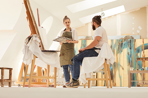 Full length portrait of female artist painting picture on easel while working in sunlit art studio with bearded man sitting on chair and posing, copy space