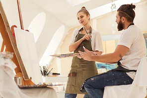 Waist up portrait of smiling young woman holding paintbrush and thinking while standing by blank canvas in art studio lit by sunlight