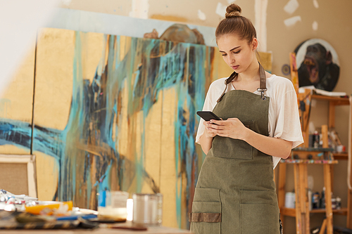Warm toned waist up portrait of blonde young woman using smartphone in art studio interior, copy space