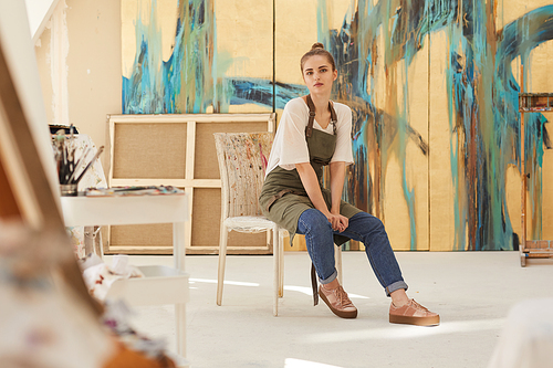 Full length portrait of contemporary female artist  while sitting on chair in art studio lit by sunlight, copy space