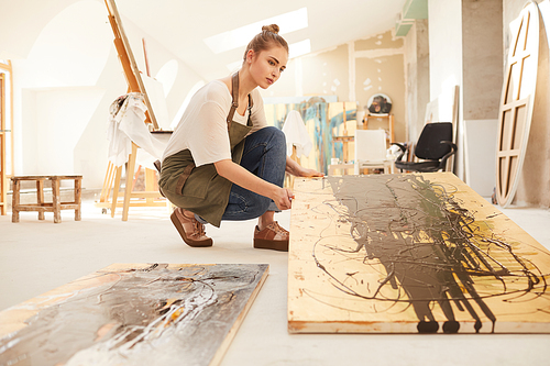 Warm-toned side view portrait of modern female artist holding abstract painting while sitting on floor in spacious art studio lit by sunlight, copy space