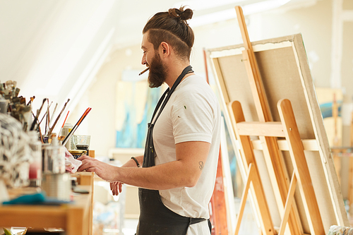 Side view portrait of contemporary bearded artist choosing tools and paintbrushes while working an art studio lit by sunlight, copy space