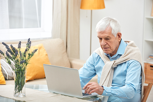 Senior man in casualwear sitting by table in front of laptop while surfing in the net and looking for online information at home