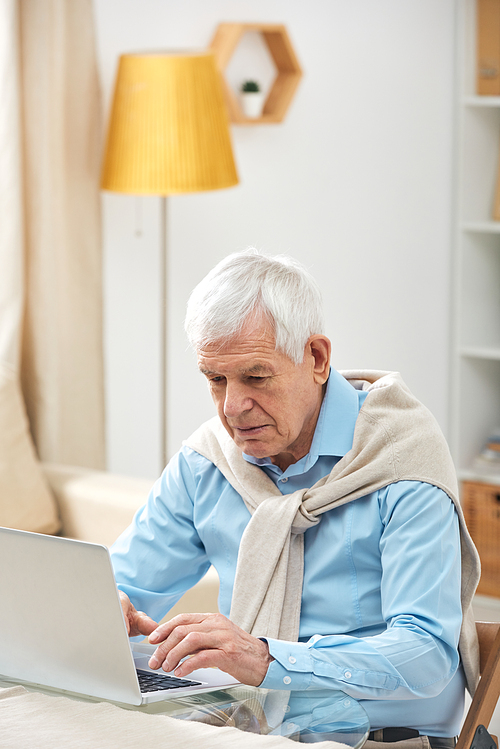 Serious busy senior man with sweated wrapped around shoulders using laptop while surfing net at home