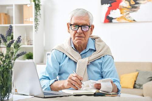 Serious senior man in eyeglasses looking through payment bills and making notes in notebook while sitting by table