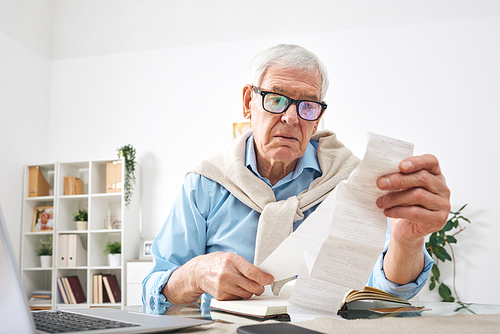 Busy senior man in eyeglasses sitting at table with laptop and diary and examining paper check at home