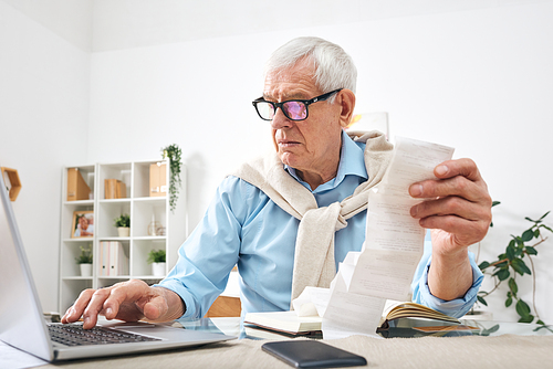 Serious senior man in eyeglasses holding payment bills while sitting by table and entering data in laptop at home