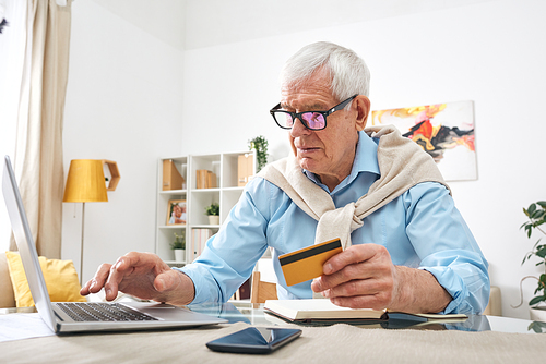 Modern elderly man with gray hair sitting at table and using laptop and debit card while buying online