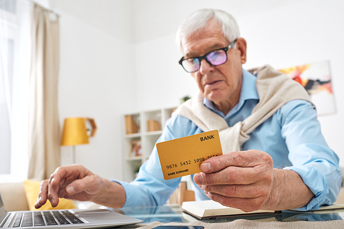 Contemporary retired man holding bank card over table while shopping online in front of laptop at home