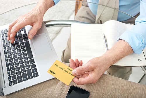 Above view of unrecognizable senior man sitting at glassy table and using laptop while paying online with credit card