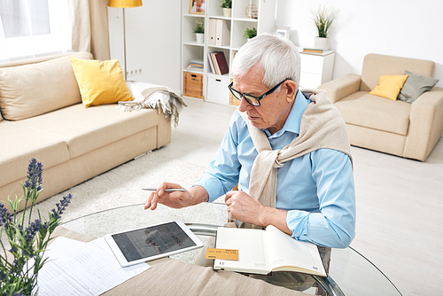 Contemporary retired man with digital tablet and bank card booking tickets online while sitting by table in his room at home