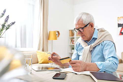 Busy elderly wrinkled man in eyeglasses using digital tablet while checking balance of debit card at home
