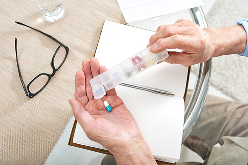 Above view of unrecognizable elderly man sitting at table with diary and using pill organizer while taking capsule