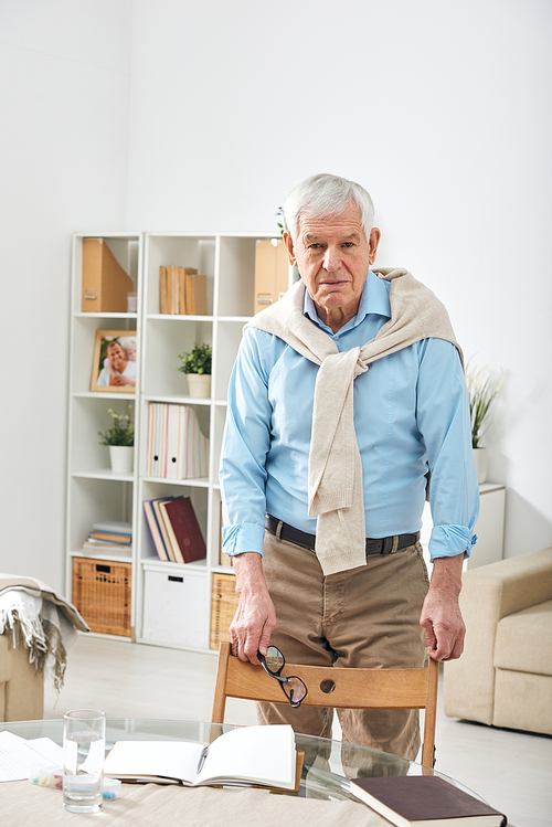 Senior retired man in casualwear looking at you while standing by chair and table with open notebook in home environment