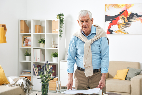 Portrait of handsome senior man in stylish outfit standing at table and viewing papers in living room
