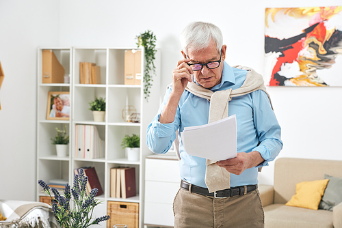 Old male pensioner in eyeglasses and casualwear looking through papers or financial documents in home environment