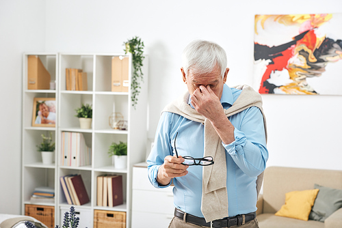 Tired senior man in blue shirt holding eyeglasses and rubbing bridge of nose while feeling eye fatigue at home