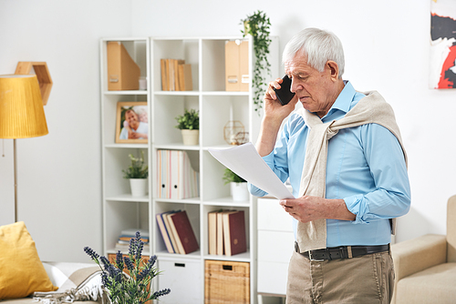 Modern senior man walking over living room and calling manager on document terms issues