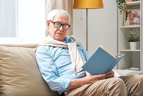 restful senior man in eyeglasses, shirt and pants looking at you while holding open book and relaxing on couch