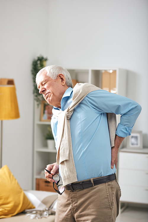 Senior man standing in living room and holding hand on lower back while feeling pain